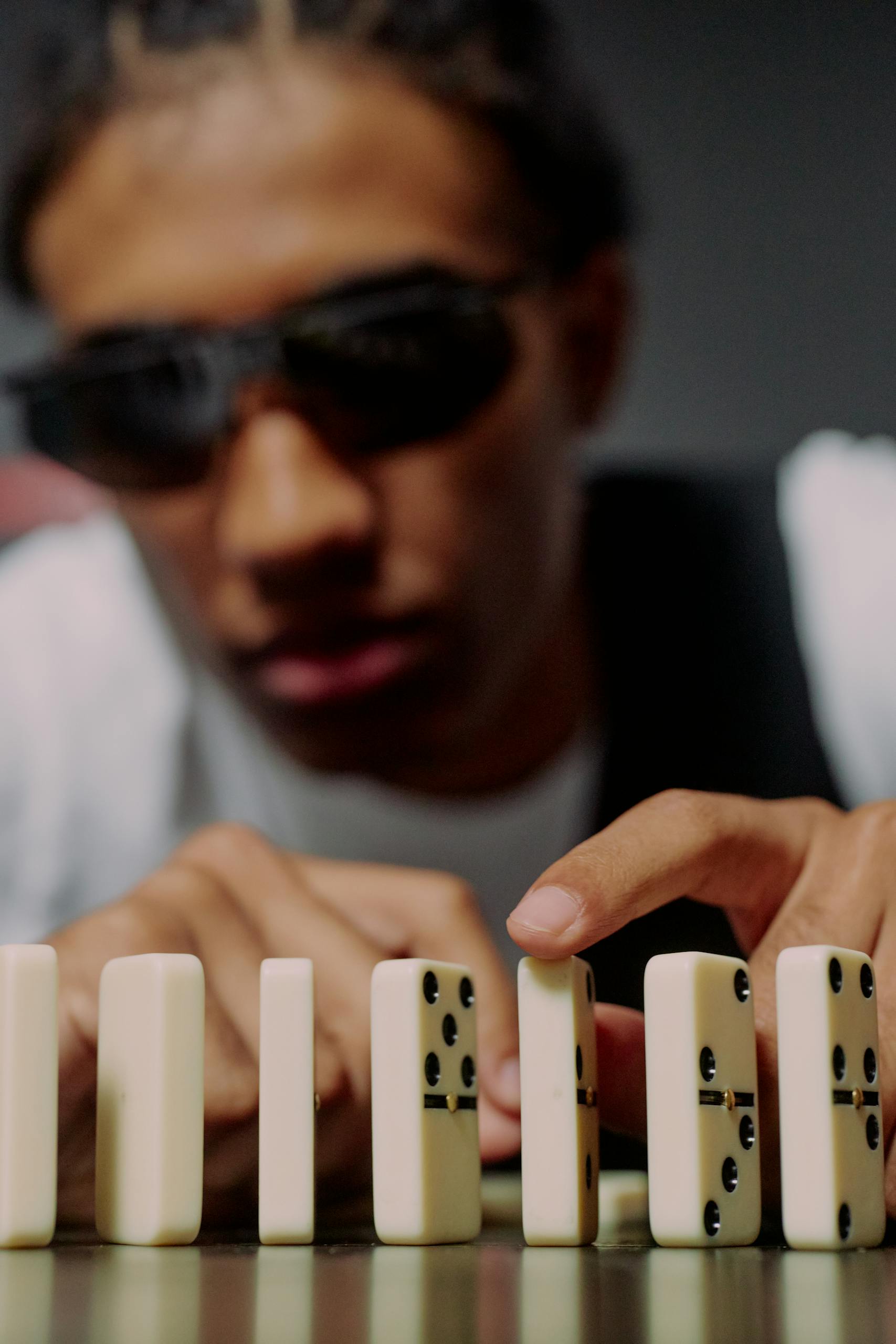 Young man wearing sunglasses arranging domino blocks indoors, focused conceptually.